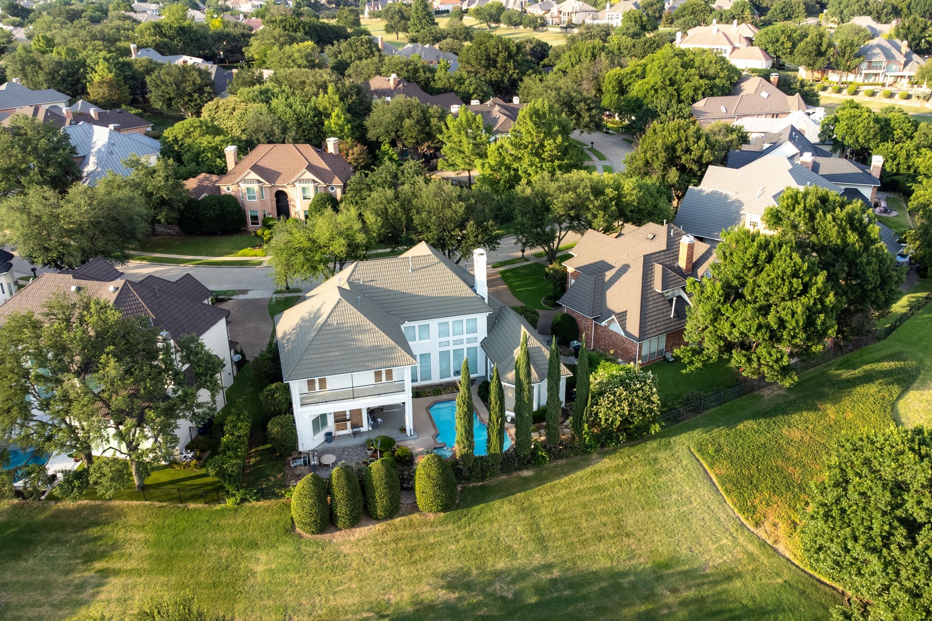 Aerial view of mansions with swimming pools surrounded by green grass and trees in the summertime.